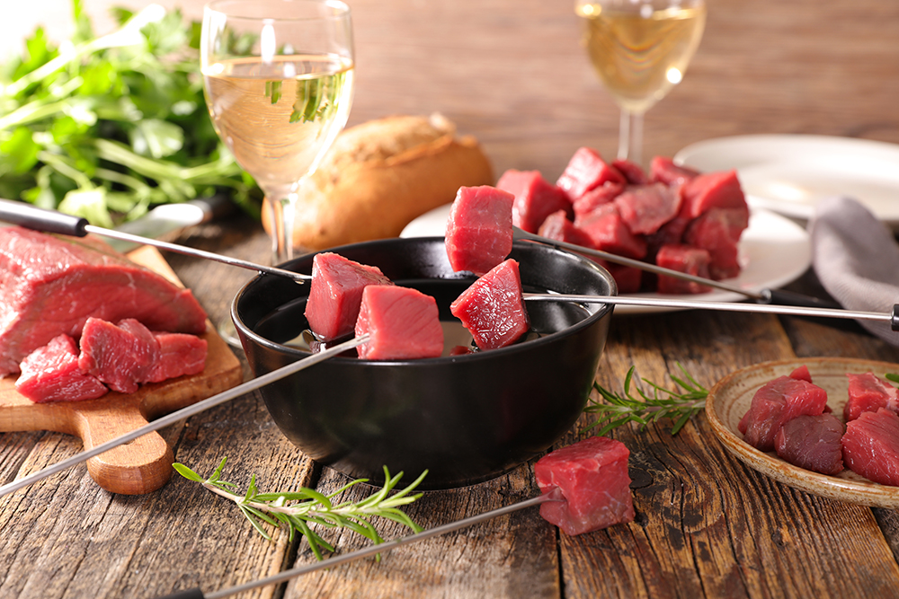 Rohes Rindfleisch in Würfeln auf Spießen über einem Topf Suppenfondue, angerichtet mit Kräutern und Weißwein im Hintergrund.