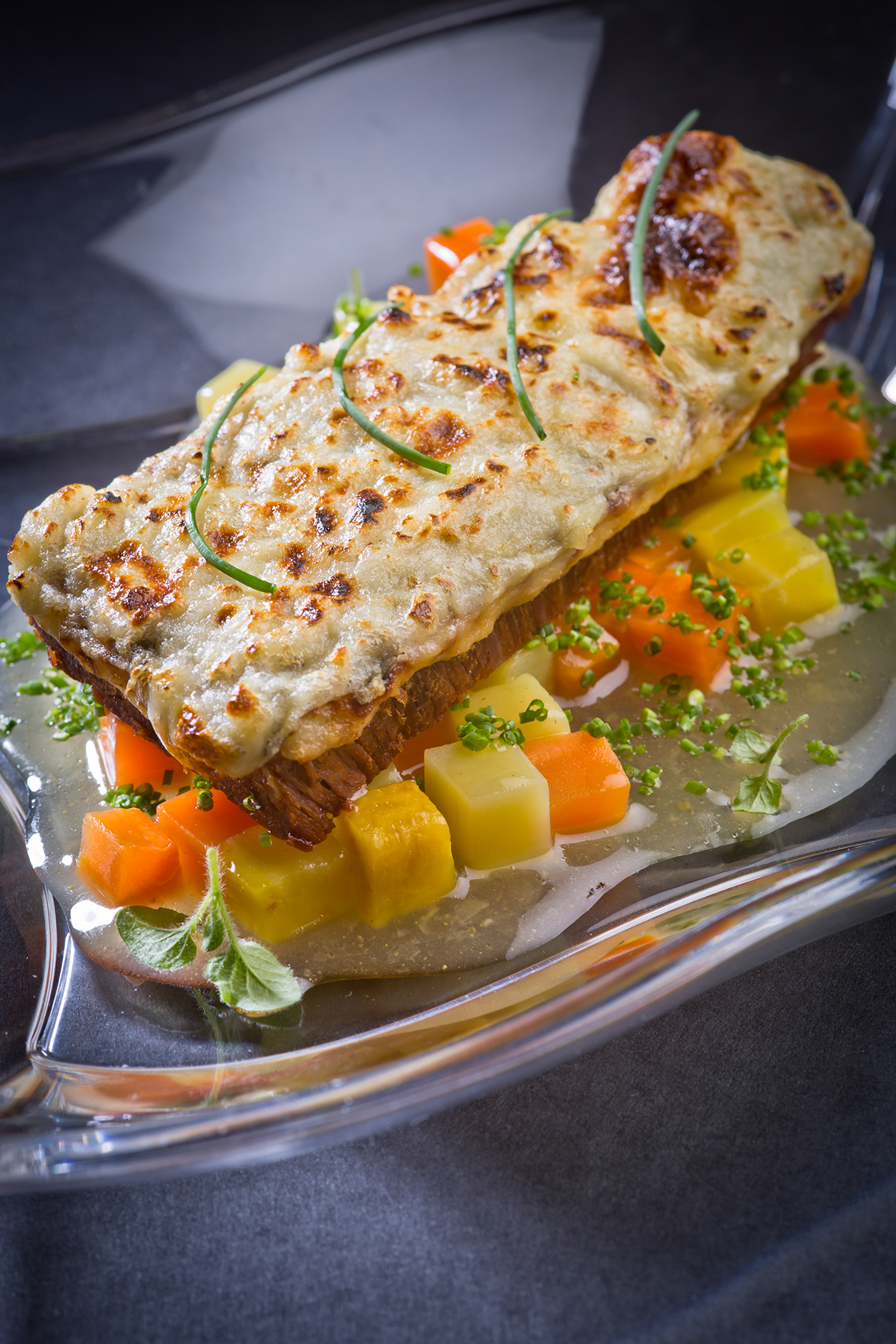 Scheibe überbackenes Rindfleisch mit Kartoffel-Karotten-Gemüsewürfeln in heller Sauce, garniert mit Schnittlauch und Kräutern.