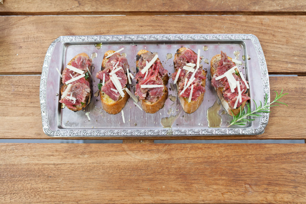 Sechs Scheiben geröstetes Brot belegt mit dünn geschnittenem, gegrilltem Rindfleisch, gehobeltem Parmesan und Kräutern auf einem verzierten Metalltablett.