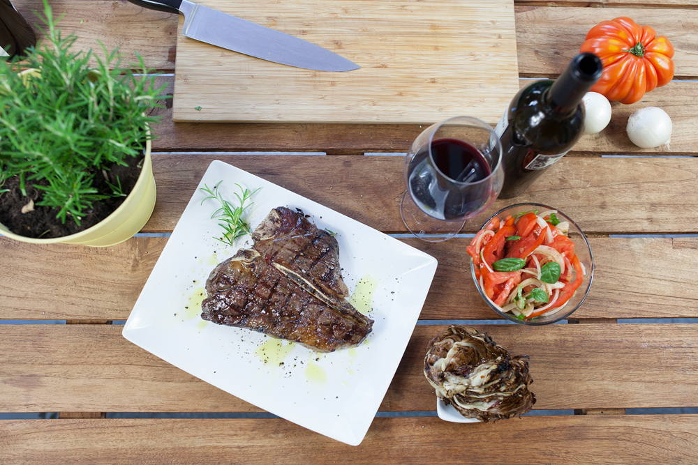Gegrilltes Porterhouse Steak auf einem weißen Teller mit Rosmarin angerichtet, daneben Tomatensalat, Radicchio und Rotwein.