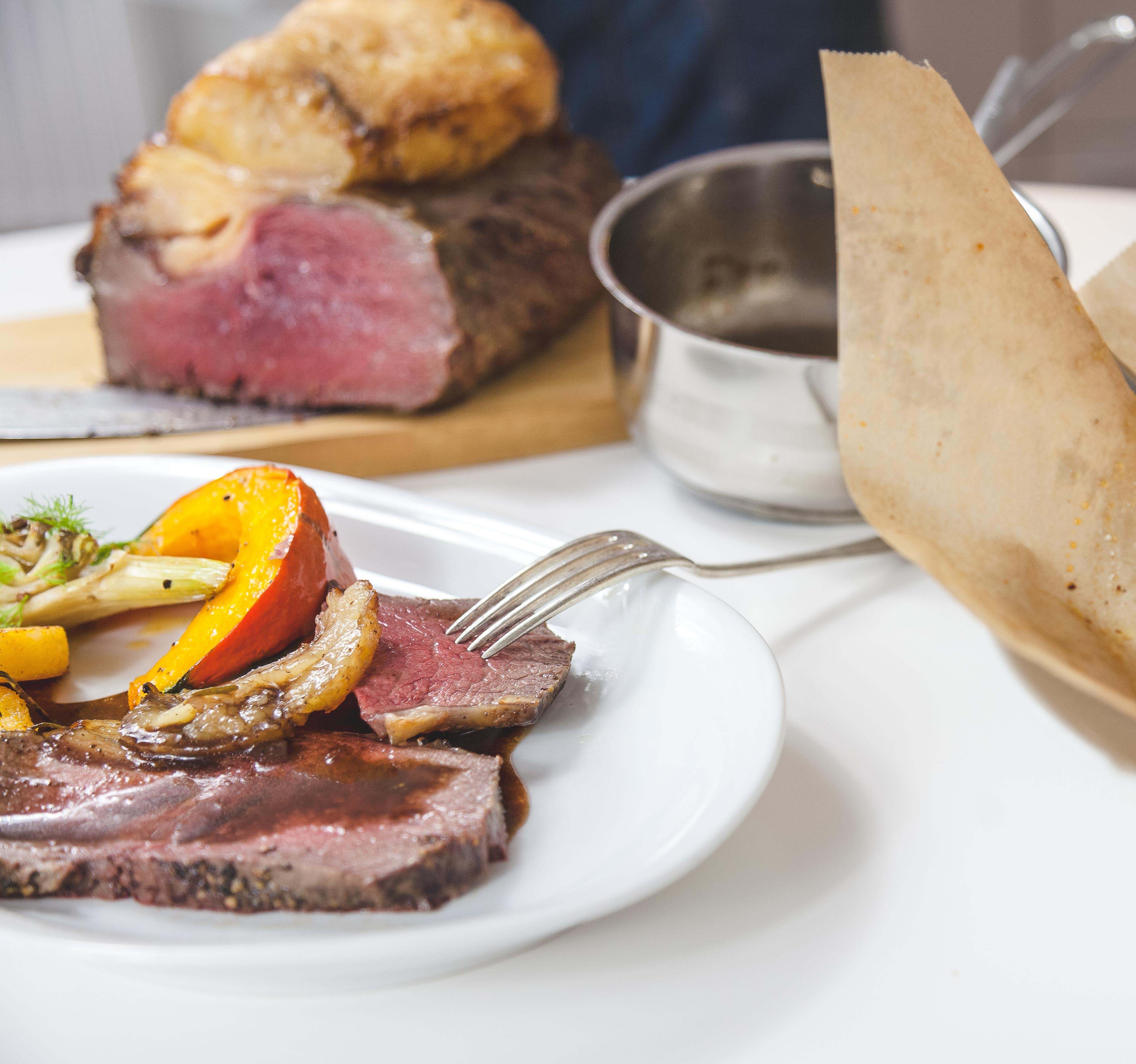 Ein angerichteter Teller mit Roastbeef und Gemüse, dahinter das restliche Roastbeef auf einem Holzbrett und eine kleine Kasserolle.