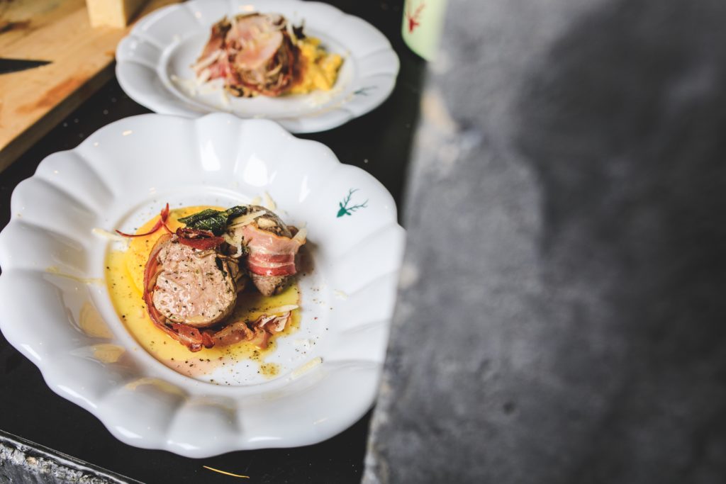Zwei angerichtete Teller mit Scheiben vom Schweinslungenbraten im Speckmantel, serviert auf cremiger Polenta und garniert mit Salbei.