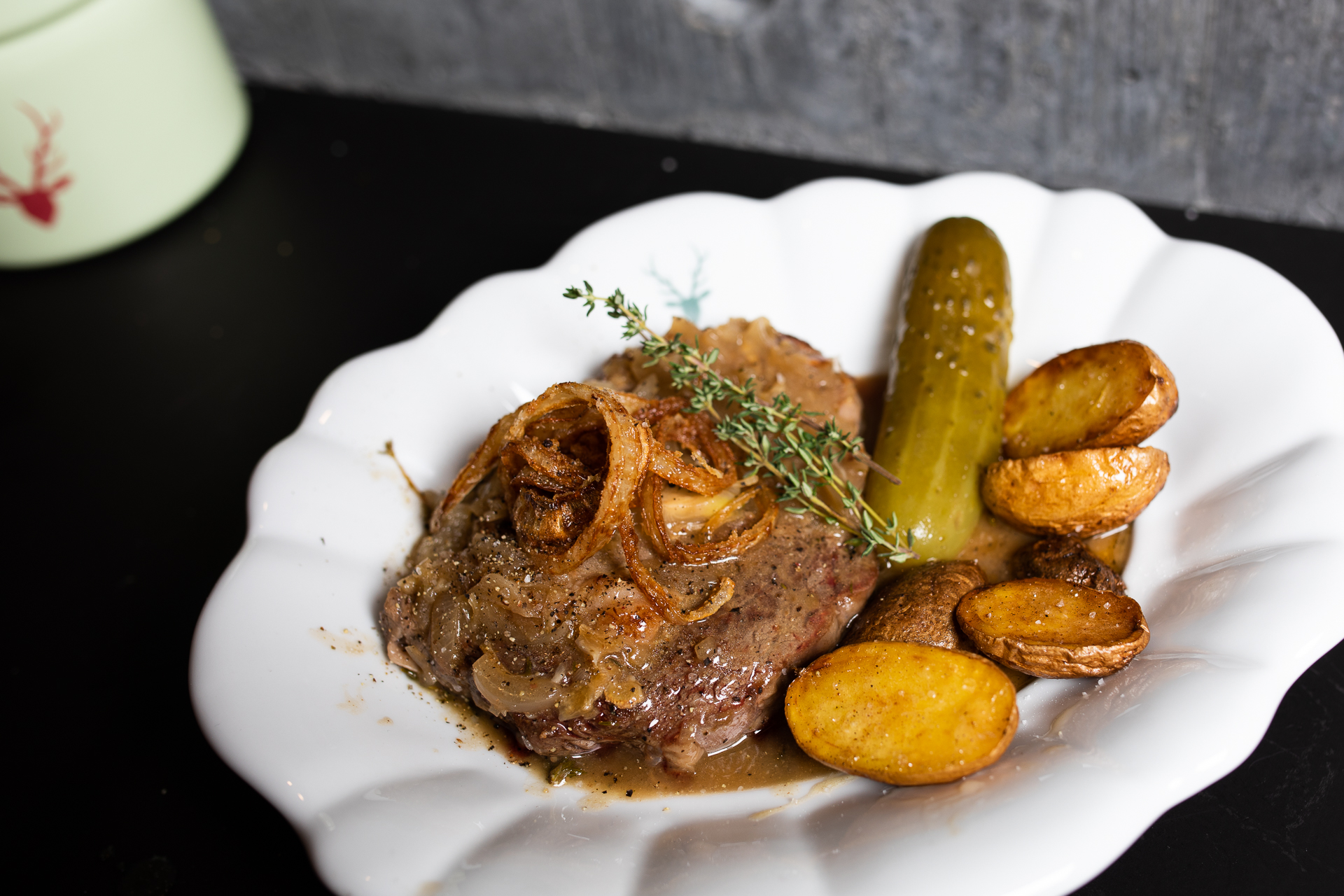 Zwiebelrostbraten auf einem weißen Teller mit Röstzwiebeln, Bratensoße, Bratkartoffeln und einer Essiggurke, garniert mit einem Thymianzweig.