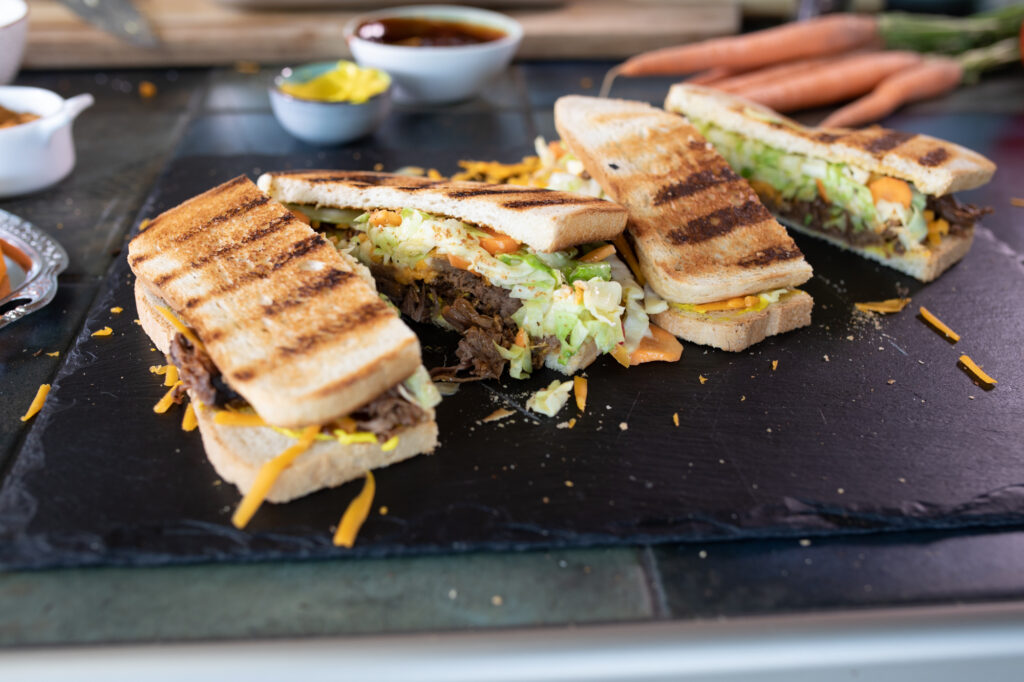 Drei gegrillte Sandwiches mit Toastbrot, gefüllt mit Pulled Beef, Käse, Salat und Karottenstreifen, angerichtet auf einer schwarzen Schieferplatte.